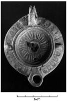 LMP-41627 - Lampe à bec en trapèze et décor d'oves SERG PRIMterre cuiteLampe moulée, anse verticale forée, à deux sillons. Réservoir circulaire à profil biconvexe. Bandeau large penché vers l'extérieur, décor d'oves doubles, deux tenons rectangulaires sur l'axe horizontal. Disque en simple cuvette limitée par trois sillons concentriques ; couronne de traits rayonnants, petite cuvette centrale. Bec en trapèze s'appuyant sur le disque, deux points au départ. Base circulaire plate limitée par un sillon, marque estampillée SERG PRIM. Engobe marron  sur toute la lampe.