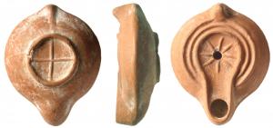 LMP-41651 - Lampe africaine à canalterre cuiteLampe moulée, anse verticale pleine. Réservoir circulaire de profil biconvexe. Deux profonds sillons larges sur le bandeau. Disque et bec moyen entourés par un bourrelet.  Huit rayons en relief aurour du trou d'alimentation. Base concave limitée par un bourrelet marquée d'une croix en relief. Engobe rouge orangé.
