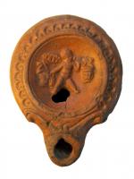 LMP-4318 - Lampe Loeschcke V S-Shaped volutes : Eros avec lanternes et raisinsterre cuiteLampe à volutes latérales de type Loeschcke V, variante égyptienne (dites à «S-shaped volute»), à petit bec rond ; médaillon concave défini par un anneau en relief, épaule légèrement convexe décorée de demi oves; orné d'un Eros ailé, dit vendangeur, portant une grappe de raisin et un vase. Base plate en forme de goutte.