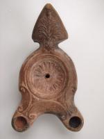 LMP-4458 - Lampe Loeschcke III à deux becs : Rosetteterre cuiteLampe à double long bec à ogive et volutes internes. Disque décoré d'une rosette. Argile noisette, engobe marron; base plate. Anse à réflecteur triangulaire avec palmette.