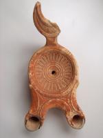 LMP-4461 - Lampe Loeschcke III : Rosetteterre cuiteLampe à double long bec à ogive et volutes internes. Disque décoré d'une rosette. Argile noisette, engobe marron; base plate. Anse à réflecteur en forme de croissant de lune.