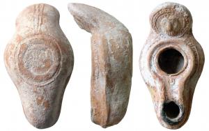 LMP-5023 - Lampe syro-palestinienneterre cuiteAnse ronde relevée portant une tête de lion de face dans une couronne de points en relief. Récipient piriforme de profil biconvexe asymétrique. Couronne de petits traits sur le bandeau. Bec long se poursuivant par une gouttière autour du grand trou  d'alimentation . Bourrelet cernant le tout.  Cercle pointé sur le bec. Base circulaire plate en anneau au léger relief. Porte une signature illisible. Trois sillons sous l'anse. Restes d'un engobe rouge foncé sur toute la lampe.