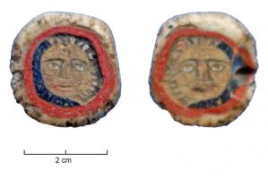 PRL-4178 - Perle polychrome  discoïdale à décor de masques verrePerle de forme discoïdale perforée dans son axe médian. Elle comporte un décor qui figure un visage sur ses deux faces. L'ensemble est entouré d'une bande bleu partiellement conservée puis d'une bande rouge et enfin d'une bande blanche. 