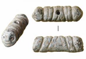 AML-3030 - AmuletteplombAmulette de forme oblongue, peut-être en forme de double phallus de part et d'autre d'une perforation centrée verticale; chaque extrémité est façonnée en forme de tête humaine schématique, mais souriante.