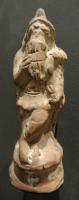 STE-4376 - Statuette : Attisterre cuiteAttis debout, en tunique et bonnet phrygien, croise les jambes dans une attitude familière d'attente; il porte à ses lèvres une flûte de Pan à 5 roseaux et tient, le long de sa jambe gauche, un attribut indéterminé (corne de pin ? corne d'abondance ?).