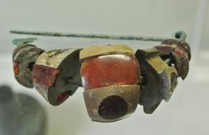 FIB-2557 - Fibule à arc compositebronzeTPQ : -625 - TAQ : -575Fibule composite : sur un arc filiforme en bronze sont enfilés des éléments formant autant de tranches juxtaposées, peut-être séparées par des éléments disparus en os ou en bois; chaque élément se compose d'un support en os, incrusté de plaquettes d'ambre. Le profil général est celui d'un arc cintré renflé (en forme de 